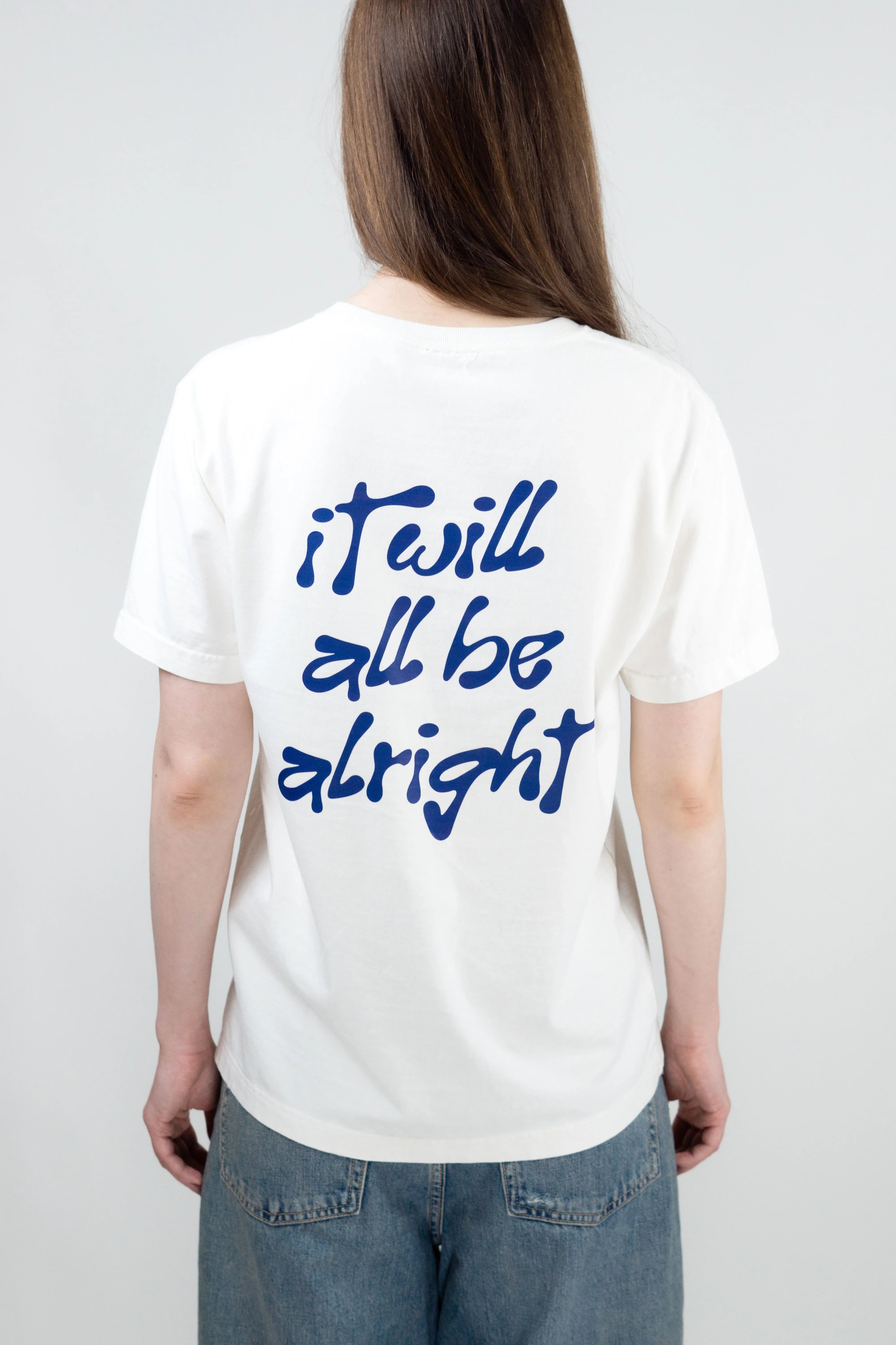 Person wearing a white t-shirt with 'it will all be alright' text on the back against a plain background