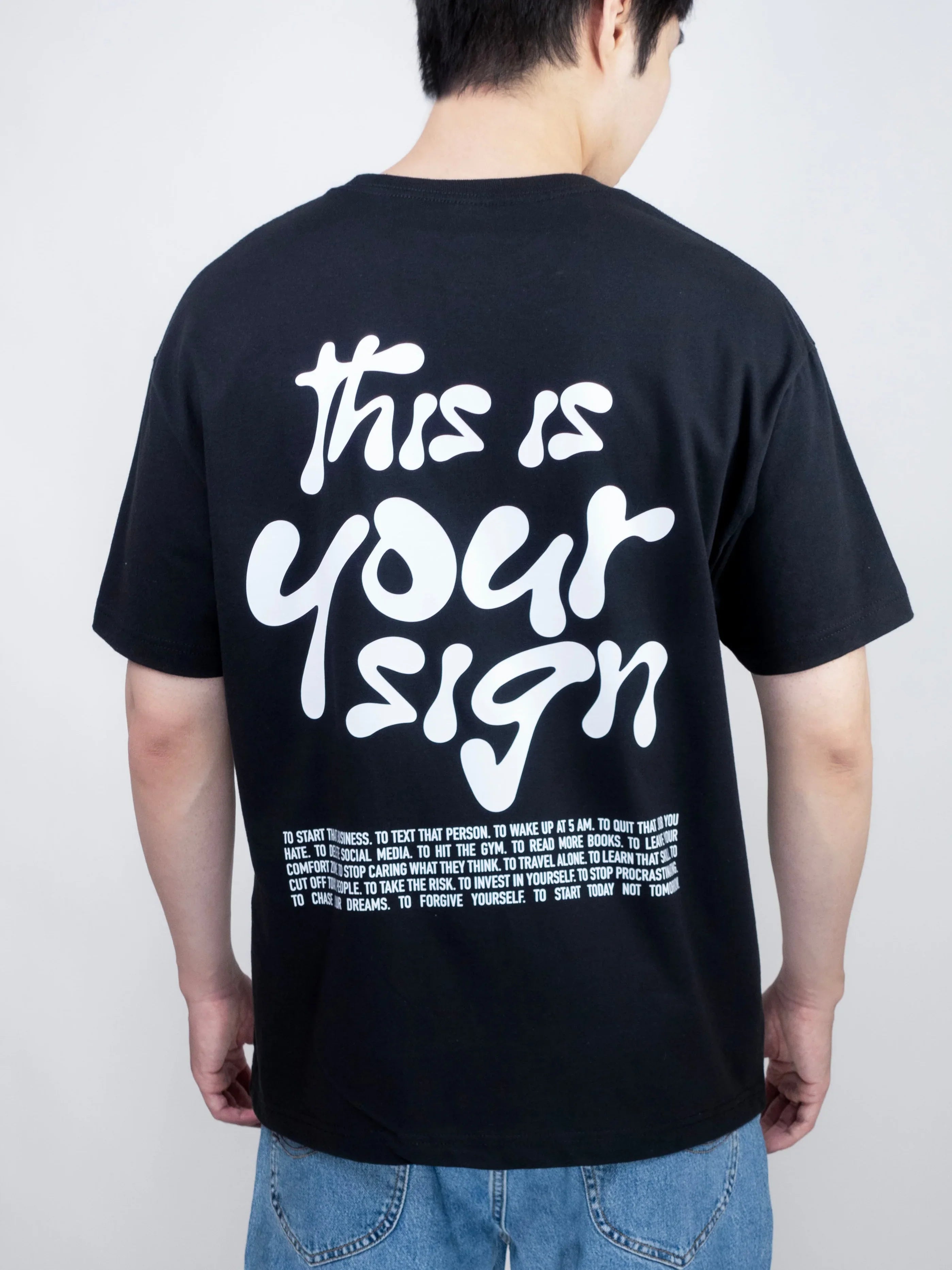 Person wearing a black t-shirt with 'this is your sign' text on the back, standing against a plain background.